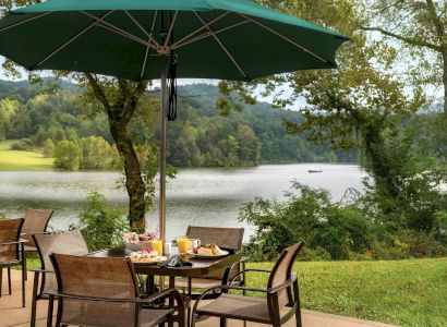An outdoor dining setup with wicker chairs and a table under a green umbrella, overlooking a serene lakeside and wooded area, with food on the table.