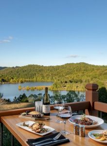 A wooden table set with plates of food, wine, and glasses overlooks a serene lake and forested hills under a clear blue sky.