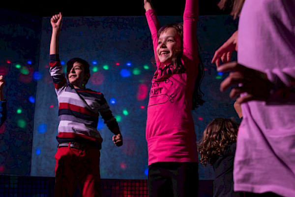 Children are joyfully dancing under colorful lights in a lively atmosphere.