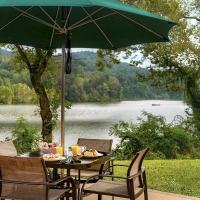 An outdoor dining area with a table, chairs, and a green umbrella overlooks a scenic lake surrounded by trees and hills, with a boat in the distance.