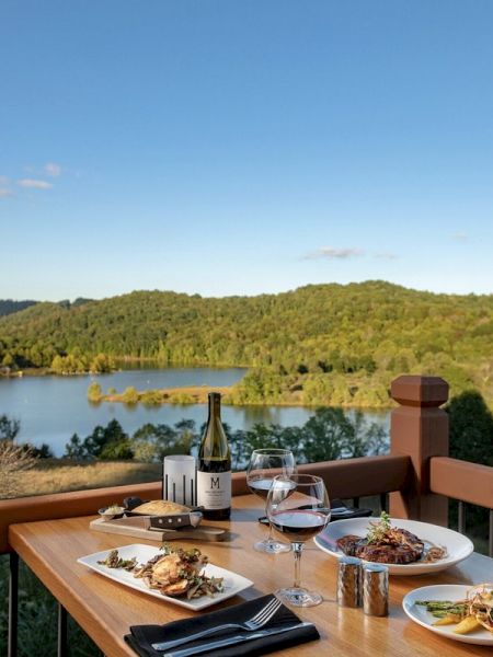 A table set for an outdoor meal with dishes, wine, and condiments, overlooking a lake and lush green hills under a clear blue sky.