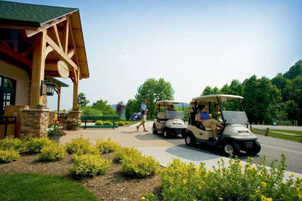The image shows two people in golf carts near a wooden building with another person carrying a golf bag, surrounded by trees and landscaping.