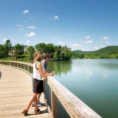 A man and woman stand on a wooden deck overlooking a tranquil lake surrounded by lush green hills and a clear blue sky.