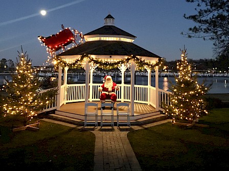 A festive gazebo decorated with lights, featuring Santa inside and Christmas trees around, set against a night sky with the moon shining.