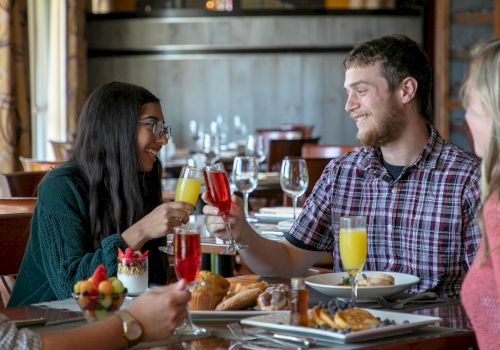 Two people are clinking glasses and smiling in a restaurant, with food spread out on the table, while two other people are visible.
