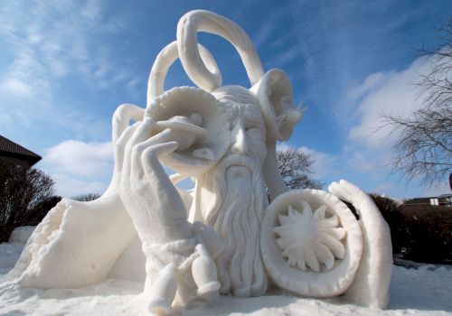 A snow sculpture depicting a bearded figure with twisted horns, holding various objects, surrounded by snow under a partly cloudy sky.