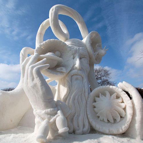 A snow sculpture depicting a bearded figure with twisted horns, holding various objects, surrounded by snow under a partly cloudy sky.