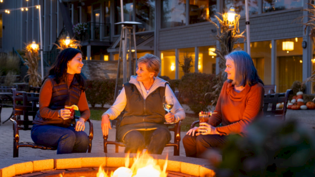 Three people sit around a fire pit, enjoying drinks, with string lights and a cozy setting in the background.