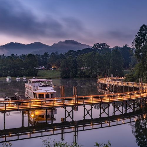 A lit wooden bridge curves over a calm body of water with a boat docked at its side, surrounded by trees and mountains in the background at dawn.