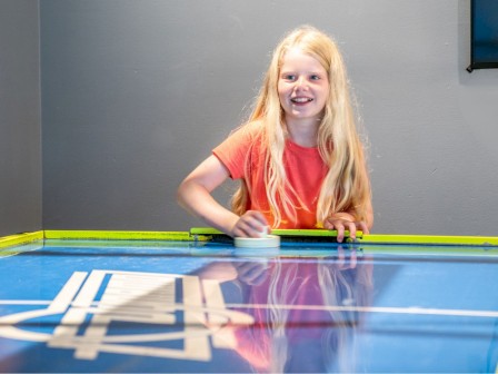 A young girl with long hair is playing air hockey, smiling, wearing an orange shirt, in a room with gray walls and a yellow-blue air hockey table.