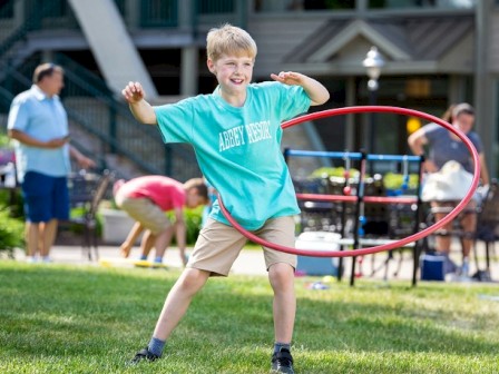 A child is playing with a hula hoop on a grassy area, with people and outdoor furniture in the background.