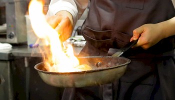 A chef flames a pan, cooking with high heat and a visible burst of fire in a busy kitchen.