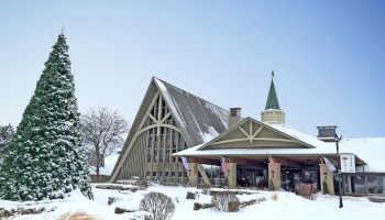 A snowy scene with a triangular church, evergreen tree, and a small building, all blanketed in white under a clear blue sky.