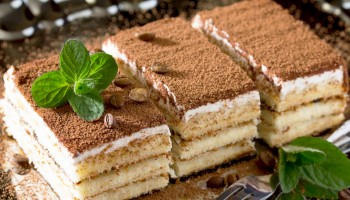A plate of layered tiramisu-like cakes with cocoa dusting, mint leaves, and coffee beans as garnish.