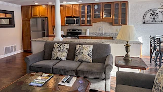 Cozy living room with gray sofa, patterned pillows, wooden coffee table, and open kitchen in background featuring stainless appliances and bar counter.