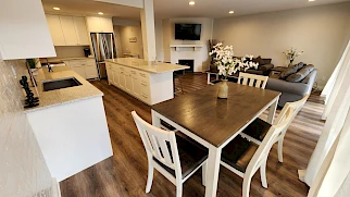 A modern open-plan kitchen and dining area with a large wooden table, white chairs, island, stainless appliances, and a cozy living room in the background, bright and inviting.
