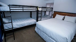 A simple hotel room with two bunk beds against the left wall and a large white king bed on the right, neutral decor, and minimal furnishings.