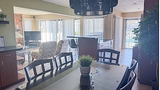 A bright open-plan dining and living area with a long dining table, black chairs, potted plant centerpiece, a blue pendant light, and large windows with vertical blinds.