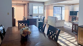 A bright open-plan living and dining area with a long wooden dining table, floral centerpiece, black chairs, a kitchen bar, and a sunny seating space with sofas and a TV.