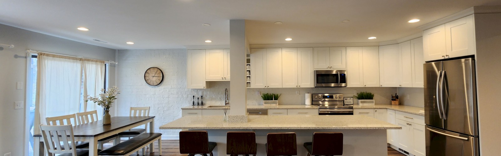 A modern kitchen with white cabinets, stainless steel appliances, an island with four stools, and a dining area with a table and chairs.