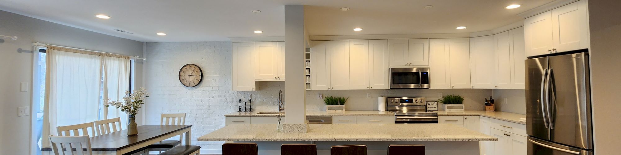 A modern kitchen with white cabinets, stainless steel appliances, an island with four stools, and a dining area with a table and chairs.