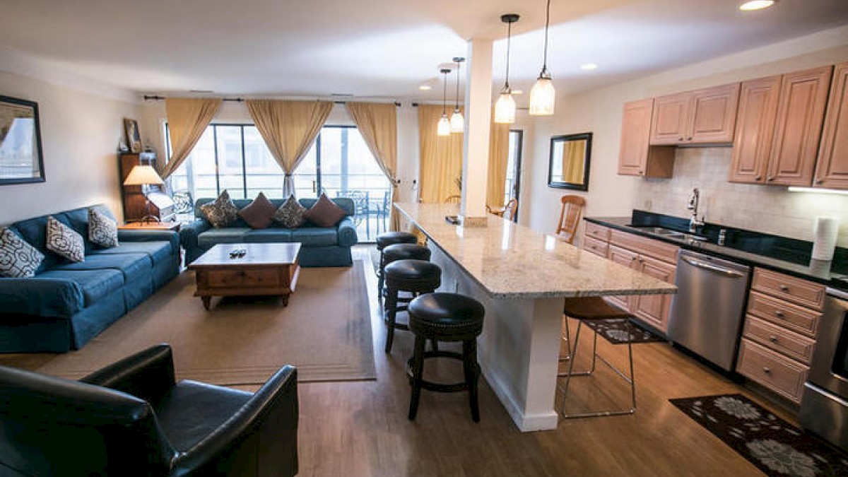 A modern living room and kitchen with a blue sectional sofa, armchair, bar stools, hanging lights, and stainless steel appliances.