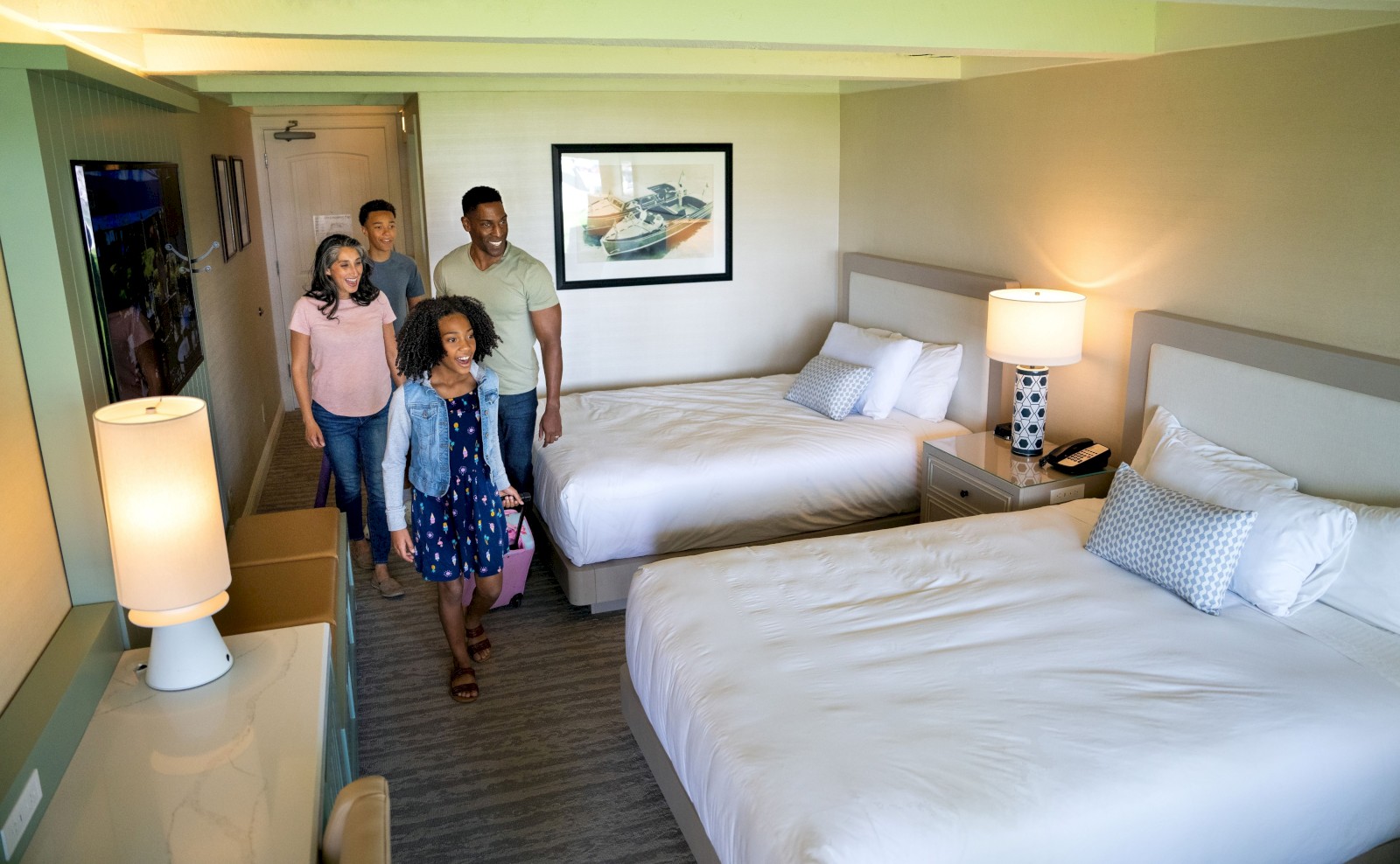 A family of four enters a bright hotel room with two beds, a dresser, and lamps. A framed picture of a ship hangs on the wall.