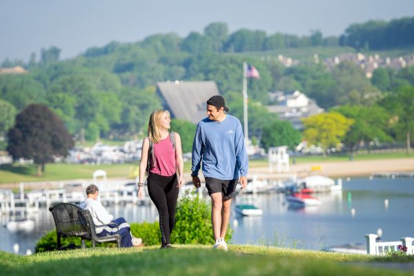 Two people are walking on a grassy hill near a waterfront, with boats, a dock, and trees in the background.