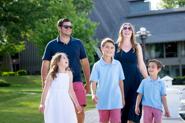 A family of five is walking outside near a building, with trees and greenery in the background. They appear to be enjoying a sunny day.