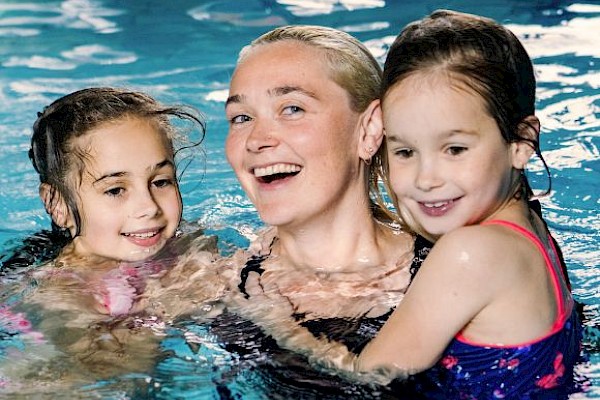 Three smiling people—an adult and two kids—enjoy a pool together, arms around each other, splashing and laughing in bright water.