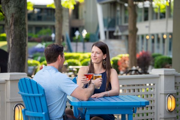 A man and a woman are sitting at a blue outdoor table, toasting drinks in a garden-like setting with buildings in the background.