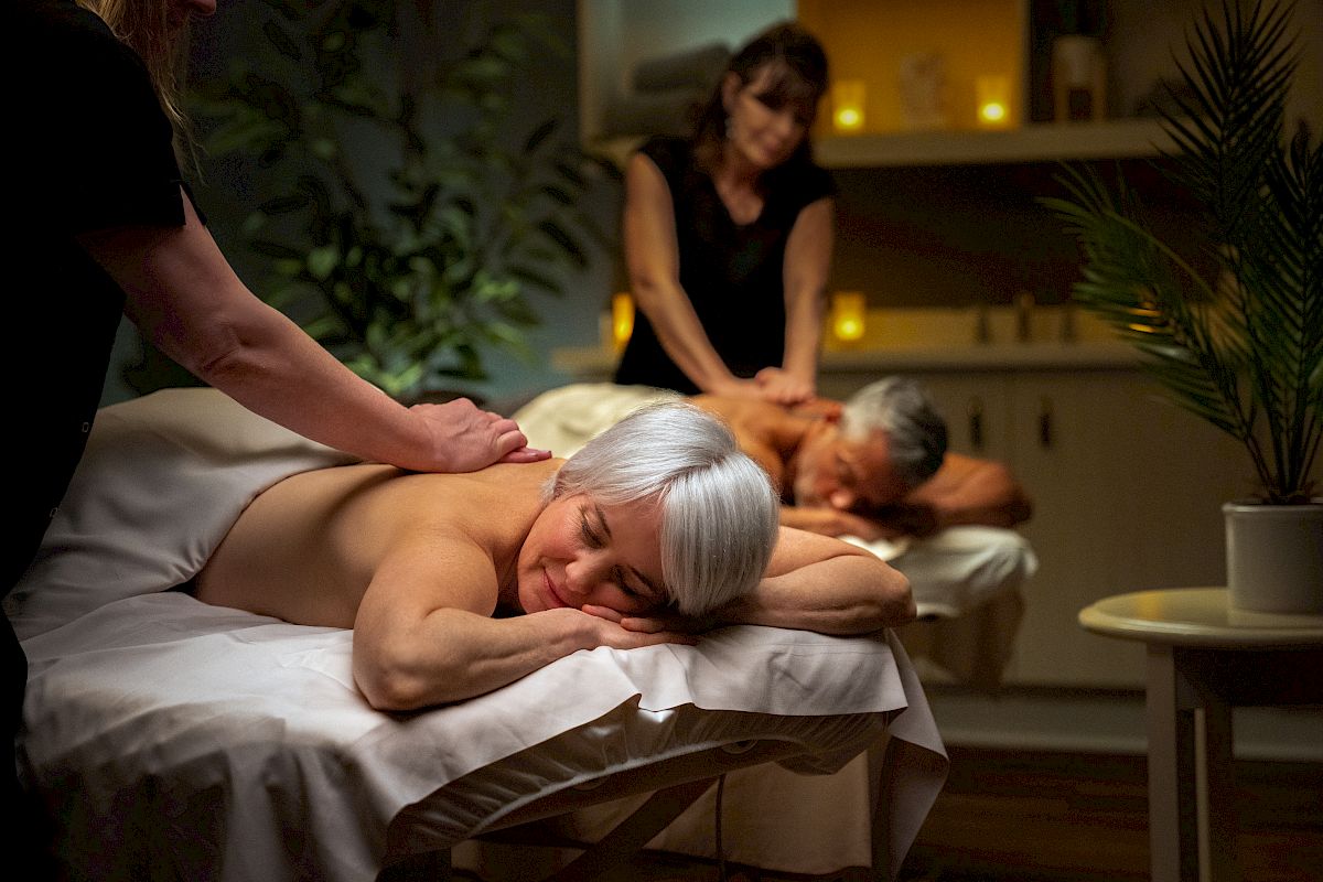 Two people receive massages on tables in a tranquil, dimly-lit room. Massage therapists attend to them, and a potted plant is nearby.