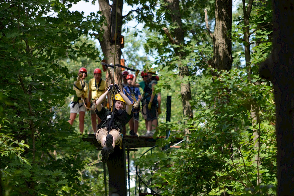 A group of people wearing helmets and harnesses stand on a suspended platform high in trees, likely enjoying a zipline or ropes course.