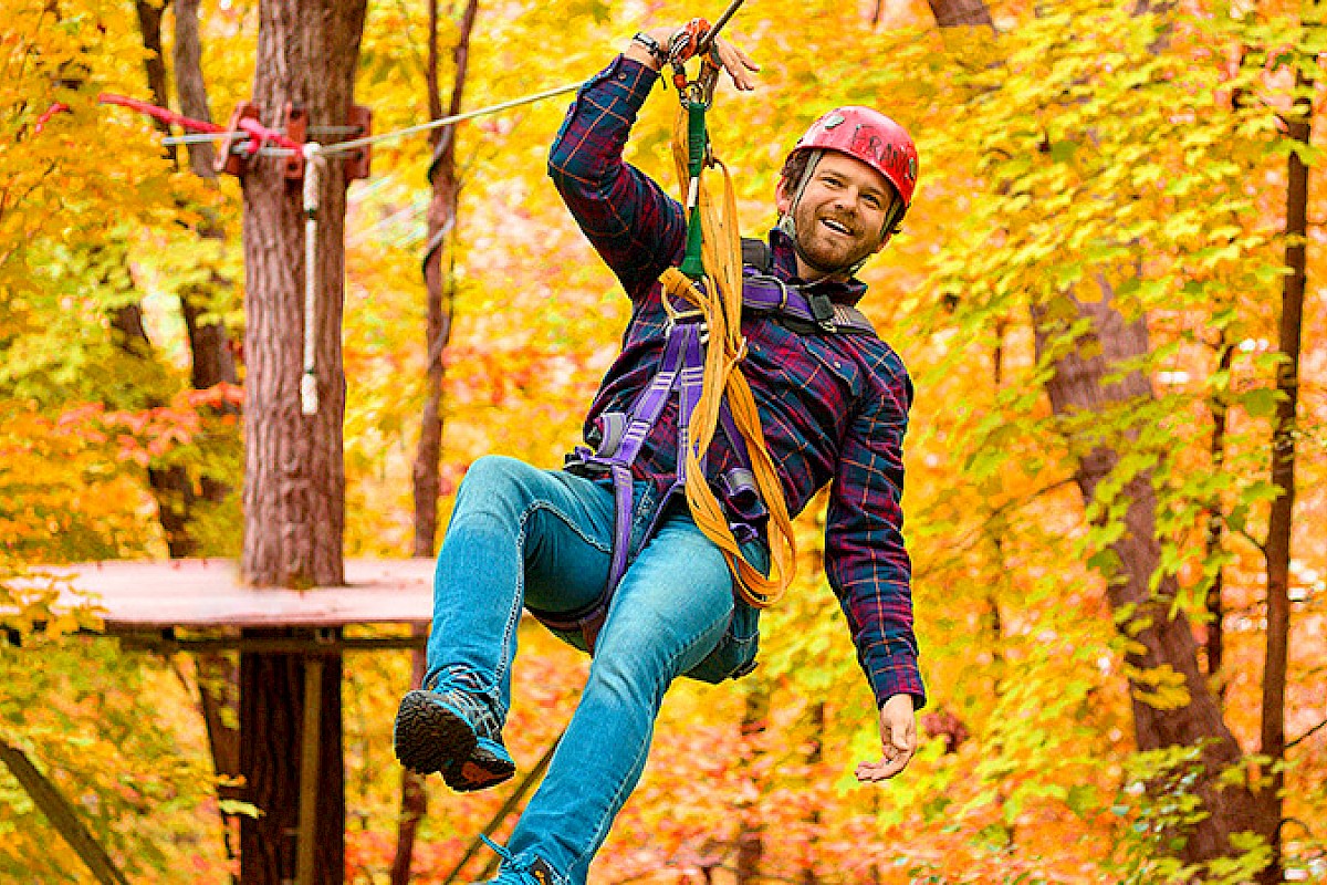 A person is zip-lining in a forest with autumn foliage, wearing a helmet and harness, smiling while holding the line.