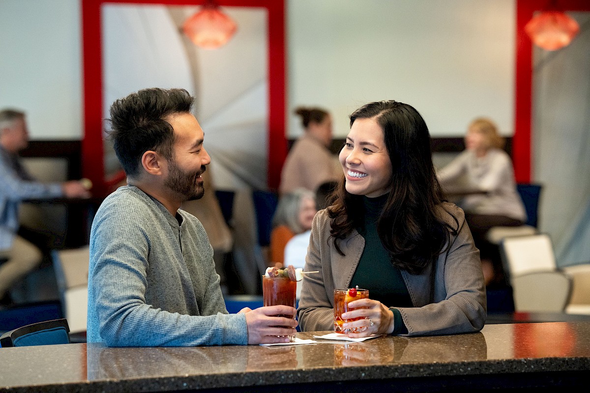 A man and woman are sitting at a bar, holding drinks and smiling at each other. Others are seated in the background.