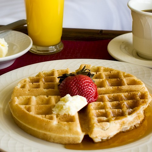 A breakfast scene: waffles with butter and syrup, strawberries, orange juice, coffee, and a cozy bed backdrop. Ending punctuation.