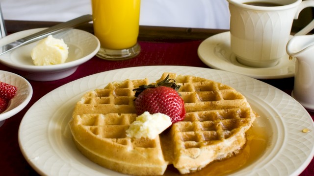 A breakfast scene: waffles with butter and syrup, strawberries, orange juice, coffee, and a cozy bed backdrop. Ending punctuation.