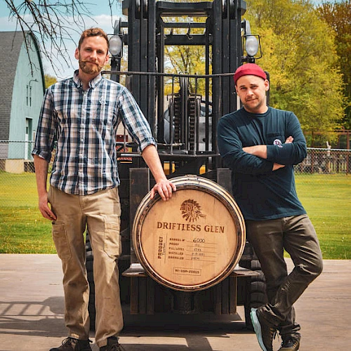 Two men stand next to a forklift holding a large barrel labeled "Driftless Glen" outdoors, with buildings and trees visible in the background.
