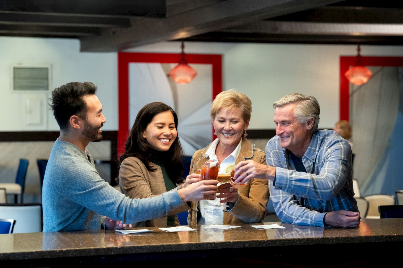 A group of four people is gathered at a bar, smiling and toasting with drinks in a cheerful atmosphere.