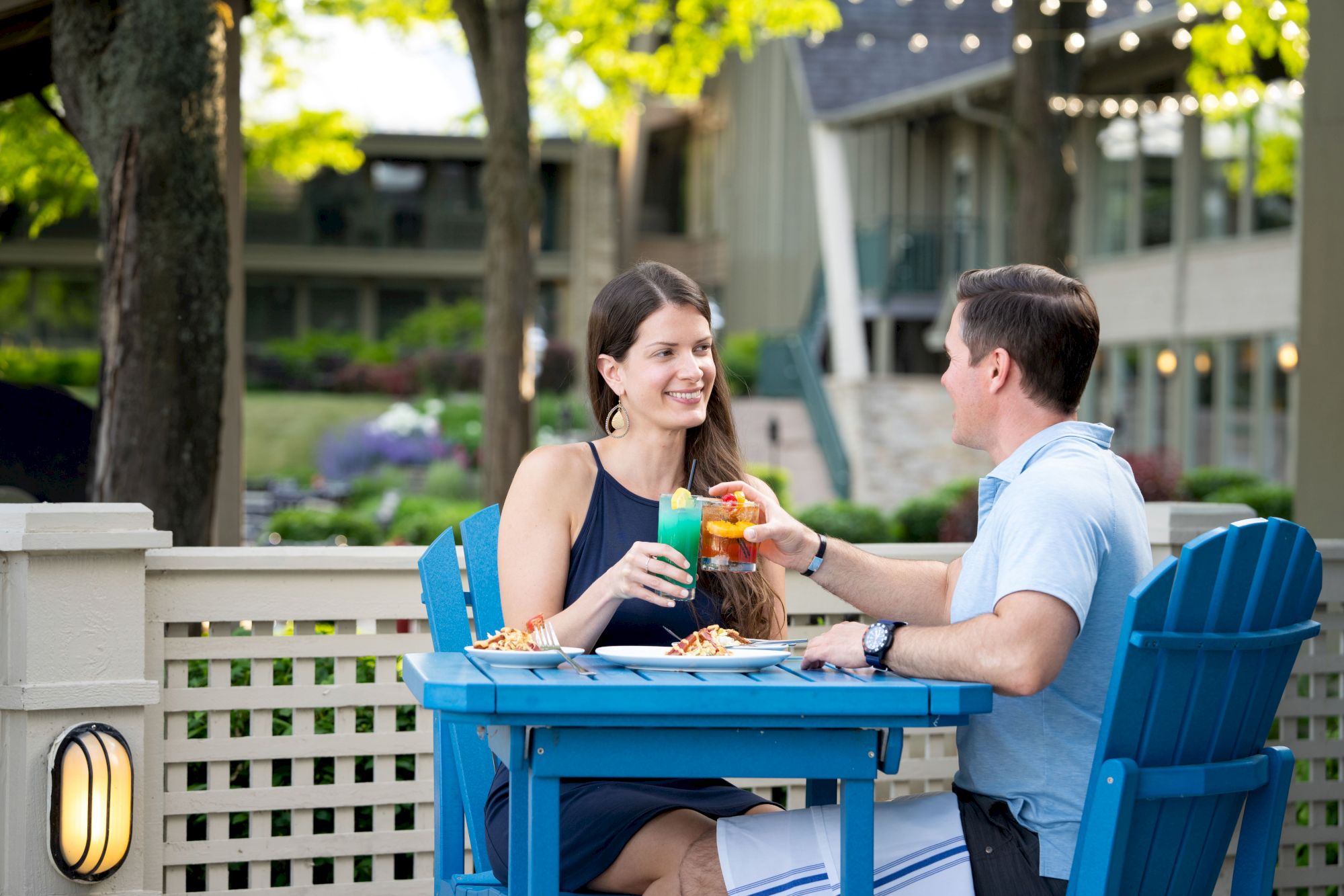 A couple sits at a blue outdoor table, toasting their drinks and enjoying a meal in a picturesque setting with trees and lights in the background.