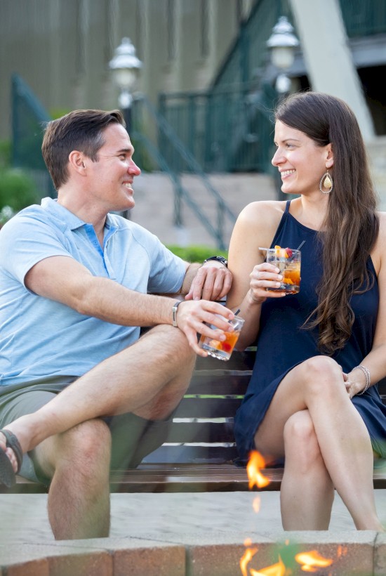 A man and woman are sitting on a bench near a fire pit, enjoying drinks outside, with greenery and buildings in the background.