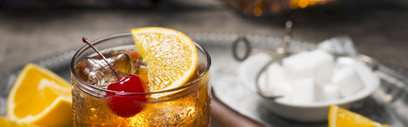 A cocktail with ice, garnished with an orange slice and cherry, sits on a tray with sugar cubes, orange slices, and a muddler.