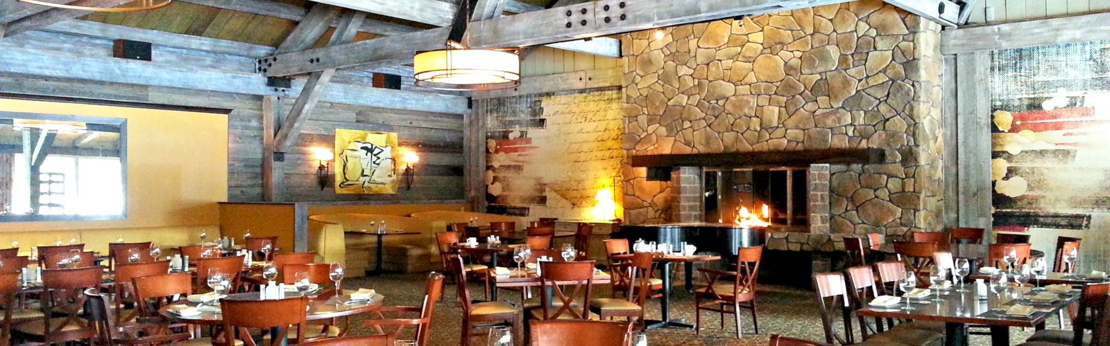 A cozy restaurant with wooden beams, stone fireplace, and neatly arranged tables set for dining. The atmosphere is warm and welcoming.