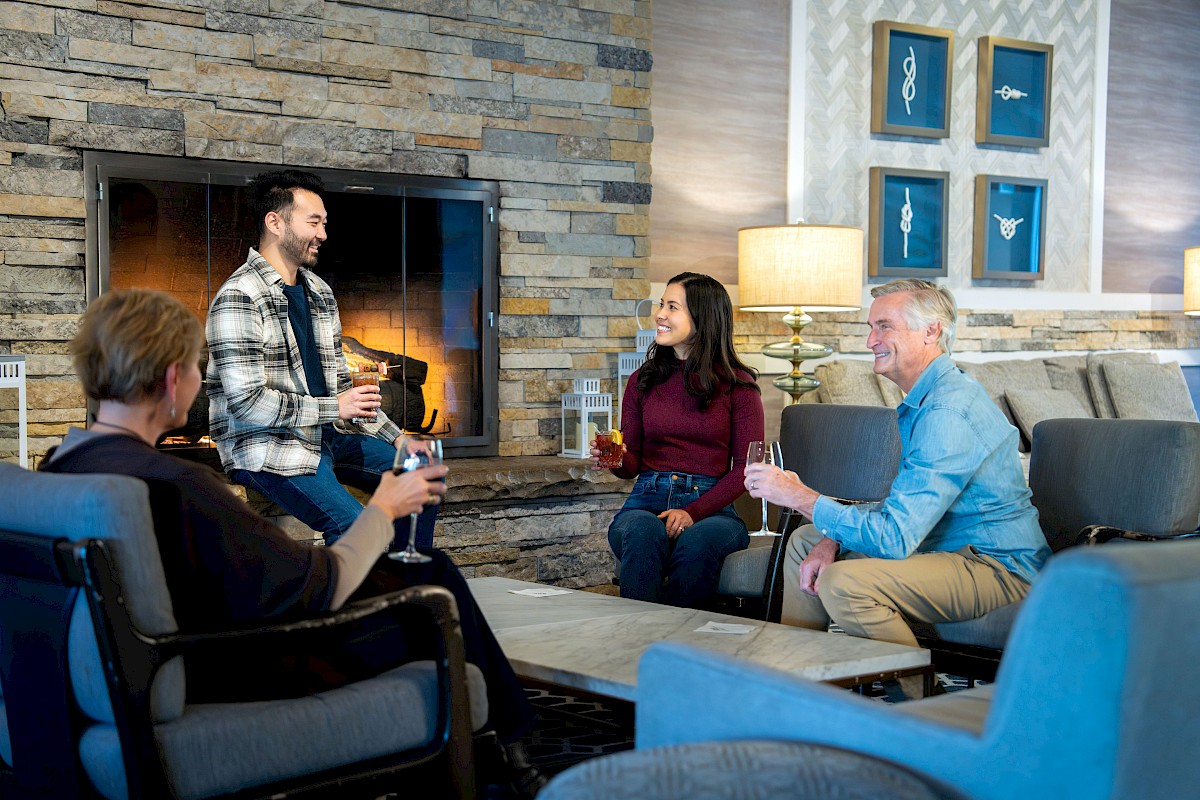 A group of four friends sits around a living room, chatting and sipping drinks by a fireplace.