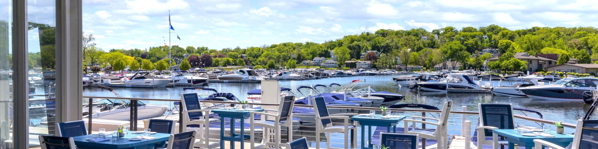 Outdoor dining area with blue tables and chairs overlooks a marina filled with boats under a partly cloudy sky.