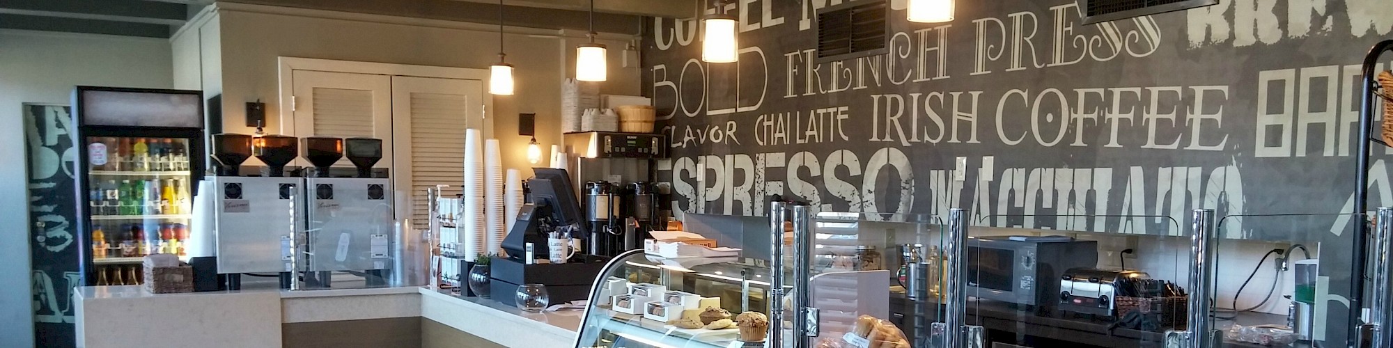 A modern coffee shop with a pastry display, beverage dispensers, refrigerated drinks, and decor featuring coffee-related words. Customers are absent.