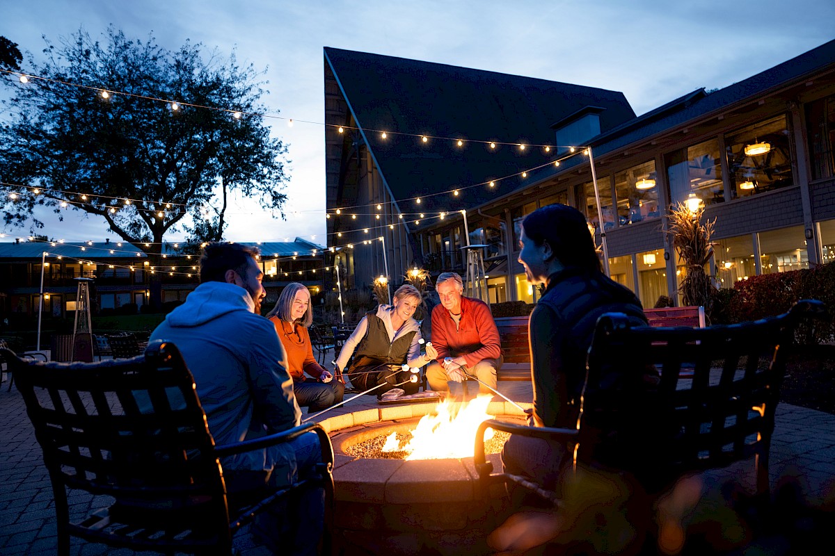 People sitting around a fire pit outdoors, surrounded by string lights in a courtyard setting, enjoying an evening gathering.