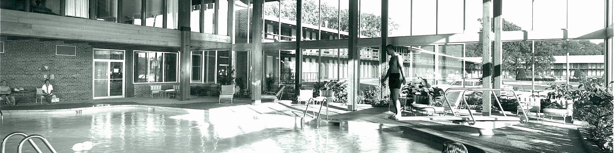 A large indoor pool area with high wooden ceilings, glass walls, and several people around the pool, including one seated on the pool's edge.