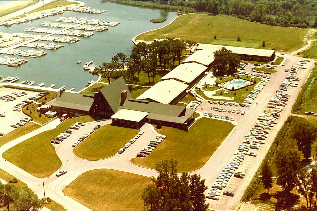 An aerial view of a marina complex with boats, buildings, numerous parked cars, and surrounding greenery.