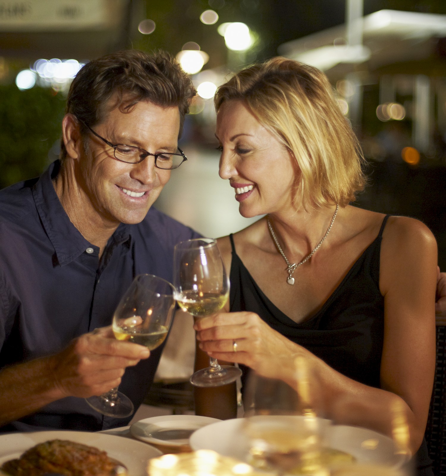A couple is dining outdoors at night, clinking wine glasses and smiling warmly, with a dimly lit ambiance.